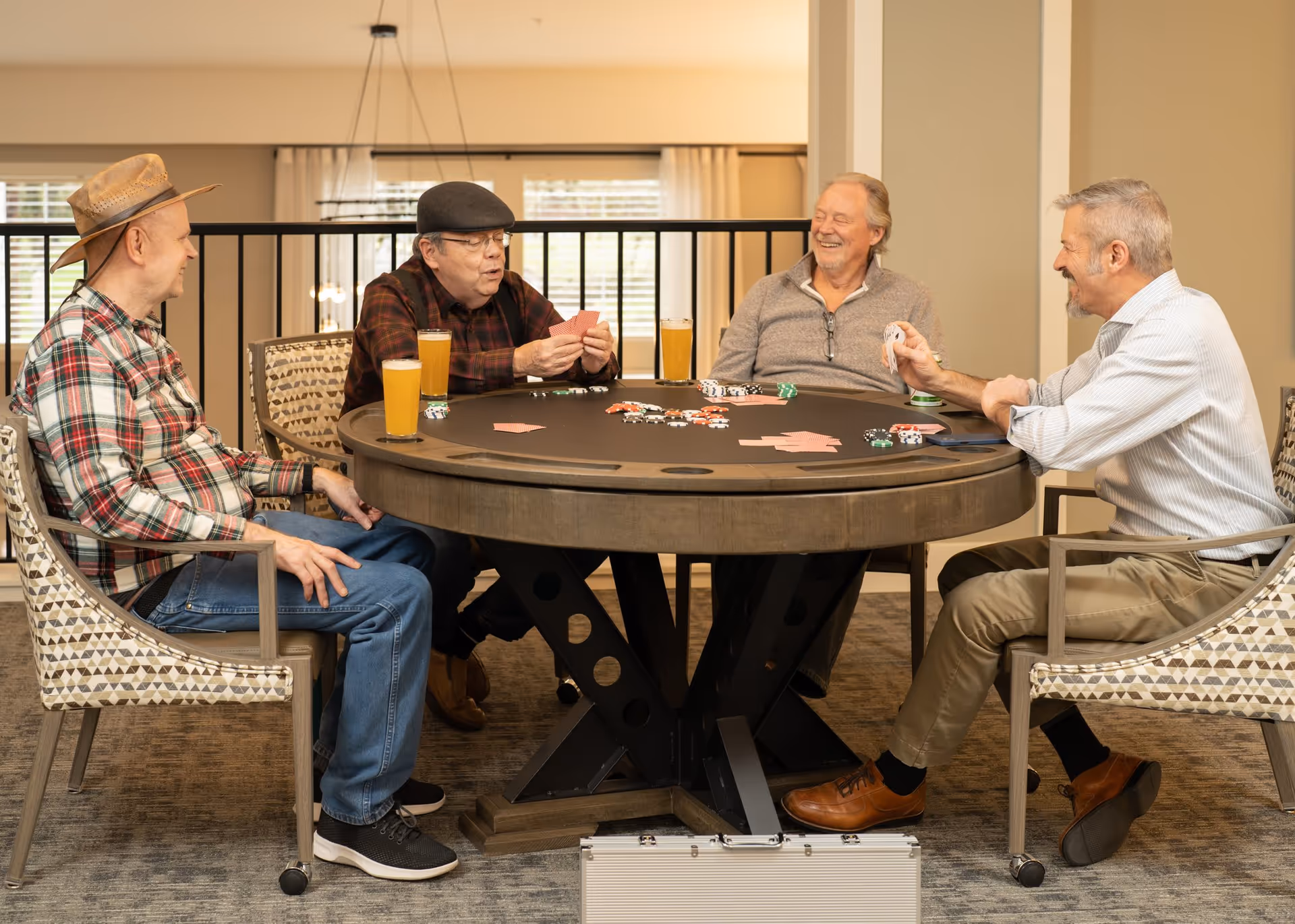 Four older men sitting around a round poker table playing cards and poker chips, with glasses of beer on the table, in a well-lit indoor common area with patterned chairs and large windows in the background.