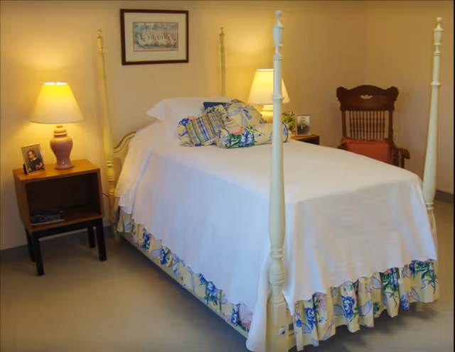 A bedroom with a four-poster bed covered in a white bedspread and floral bed skirt. The bed has several decorative pillows. On the left side of the bed is a wooden nightstand with a purple lamp and a framed photo. On the right side, there is another lamp and a small framed picture. A wooden chair with a red cushion is placed against the wall in the background. A framed picture hangs on the wall above the bed.