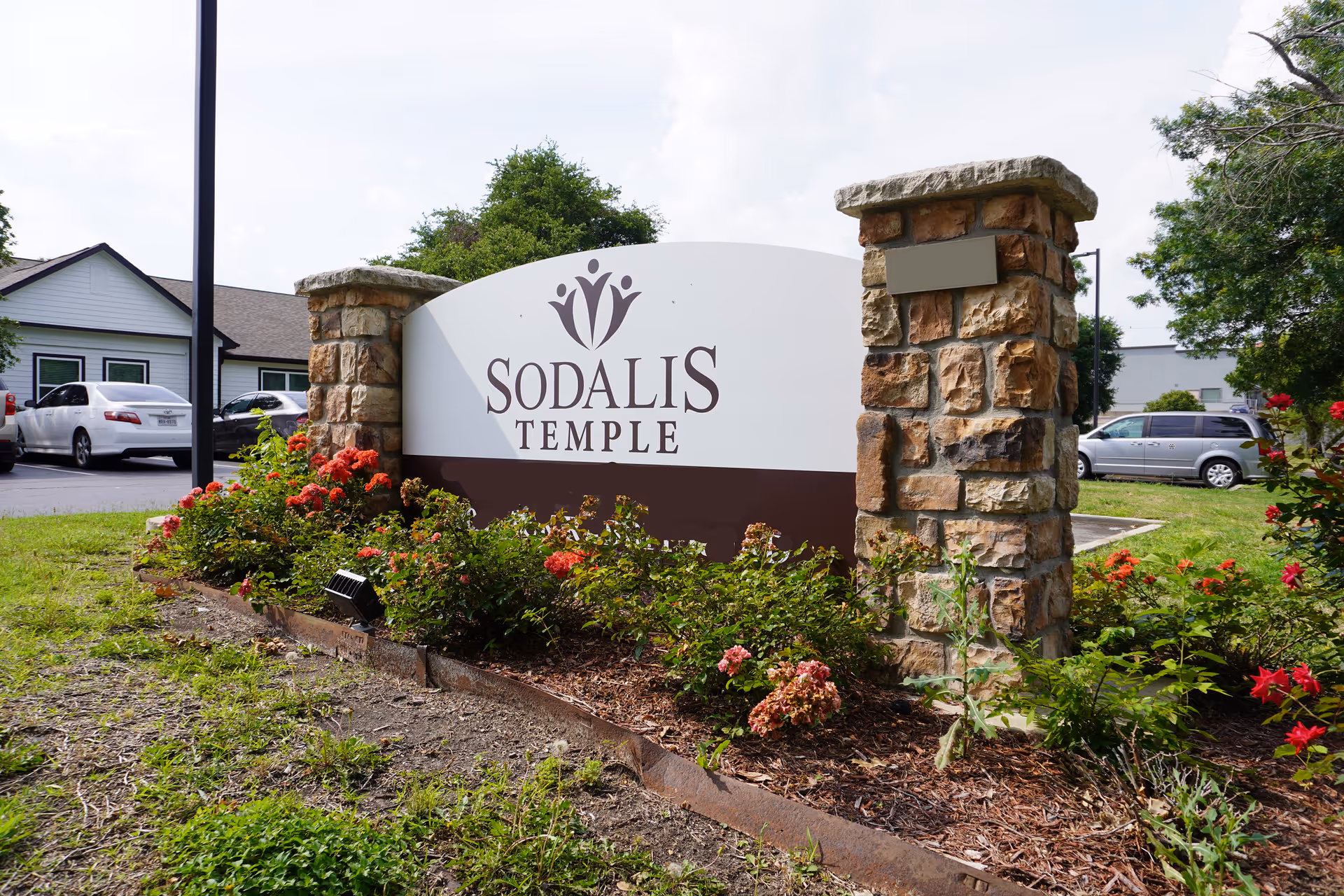Stone-pillared entrance sign reading "Sodalis Temple" surrounded by flowers and landscaping with buildings and parked cars in the background.
