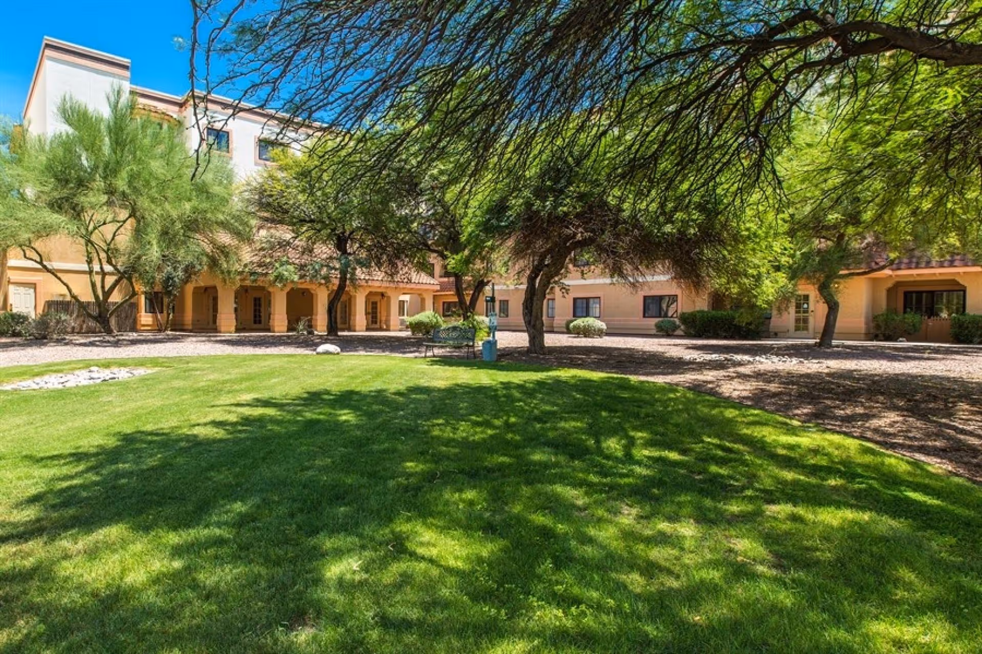 A sunny outdoor courtyard area with green grass, trees providing shade, and a bench. The background shows a multi-story building with a beige exterior and a tiled roof, featuring several windows and doorways.