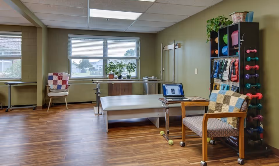 A therapy or exercise room with wooden flooring and green walls. The room features a padded therapy table, parallel bars for walking exercises, a chair with a patchwork quilt, a small table with a laptop, and a shelving unit holding colorful exercise weights and equipment. Two large windows provide natural light and a view of the outside.