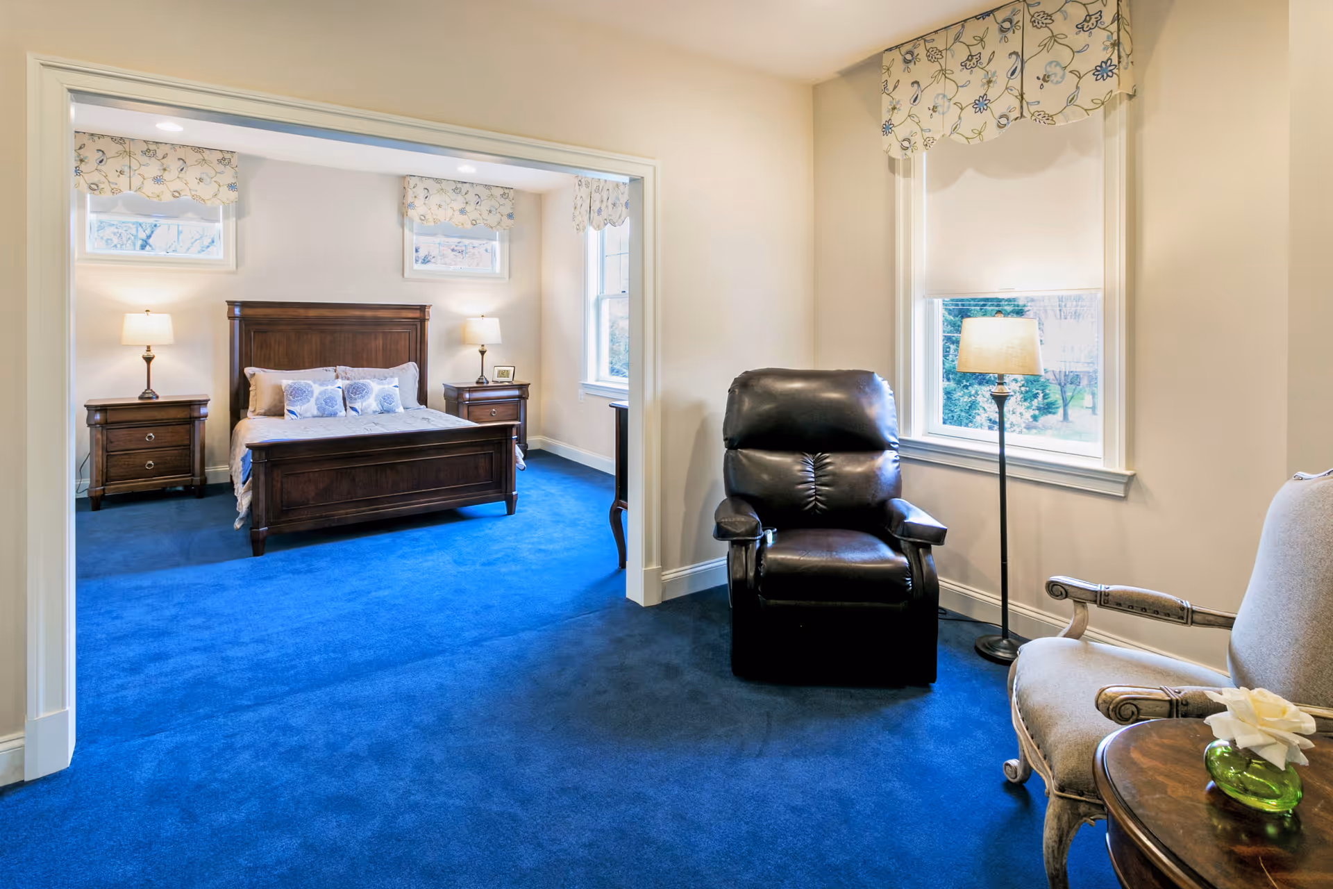 A cozy senior living bedroom with a dark wooden bed frame, two matching nightstands with lamps, and floral curtains on the windows. The room has blue carpet and an adjacent sitting area with a black leather recliner, a beige armchair, a small round wooden table with a green vase and white flower, and a floor lamp near a window.