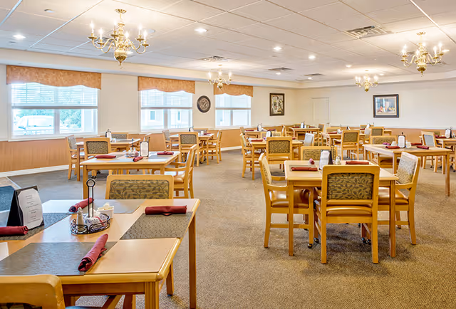 Spacious senior living dining room with multiple wooden tables and chairs set with napkins and chandeliers.