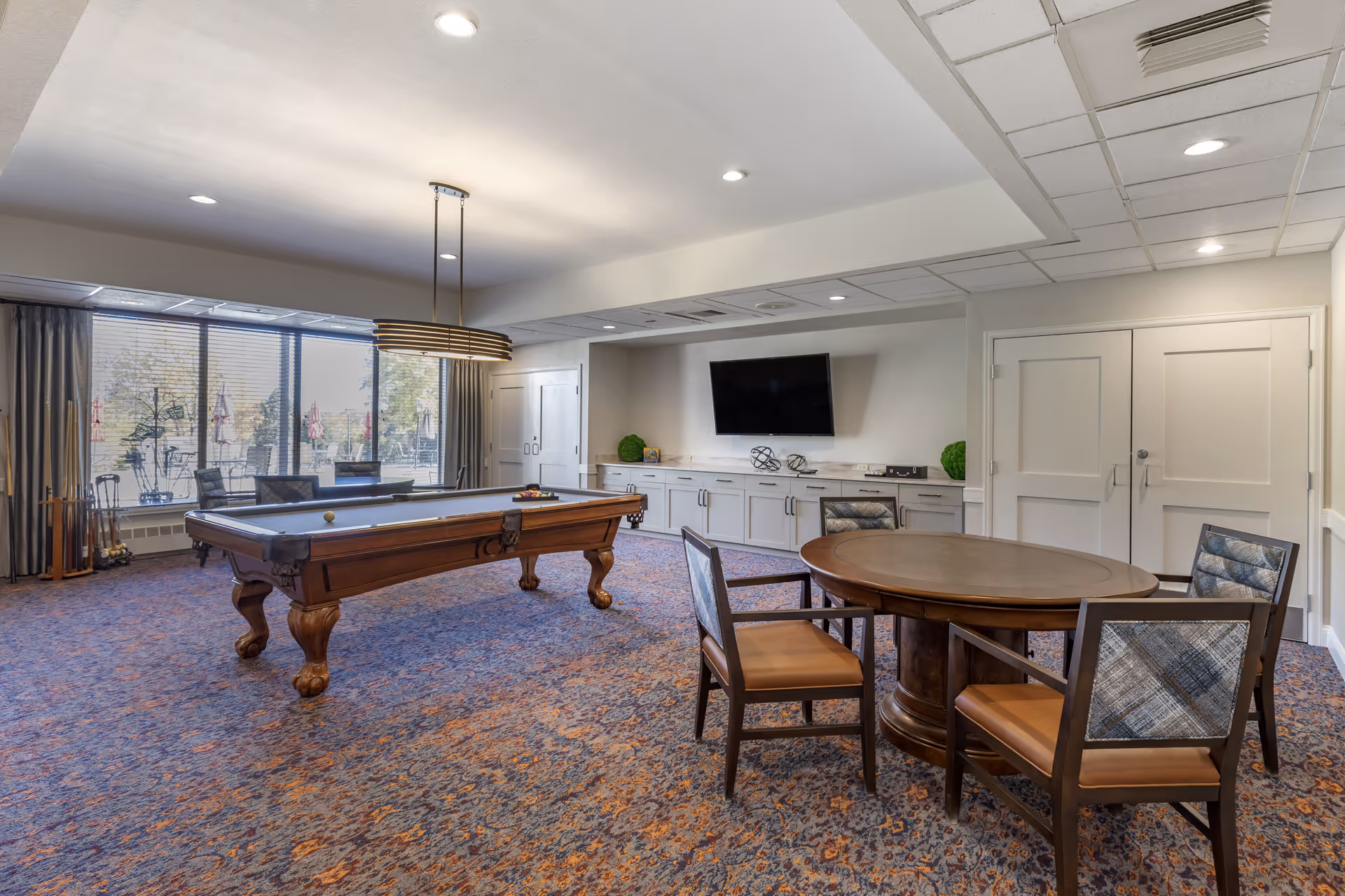 A spacious recreational room with a pool table in the center, a round wooden table with four chairs, a wall-mounted flat screen TV, and large windows with a view of an outdoor patio area. The room has patterned carpet flooring and recessed ceiling lights.