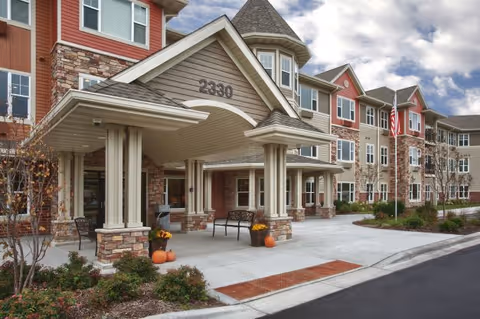 Front exterior view of a senior living facility building with a covered entrance, stone and siding facade, benches, potted plants with pumpkins, and an American flag on a flagpole. The building number 2330 is visible above the entrance.