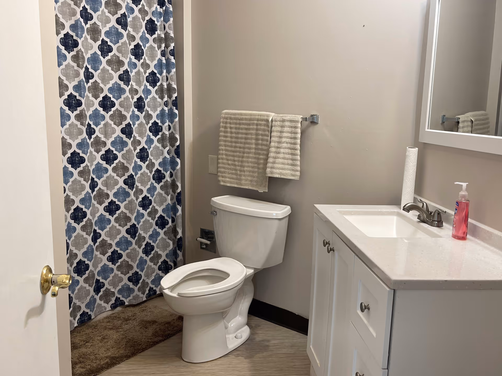 A bathroom with a white toilet, a white vanity with a sink and faucet, a mirror above the vanity, a paper towel roll and a pink soap dispenser on the countertop. There is a towel rack on the wall with two beige striped towels. A shower curtain with a blue, gray, and white geometric pattern is visible next to the toilet. The floor has a light wood-like finish and a brown bath mat is placed in front of the shower.