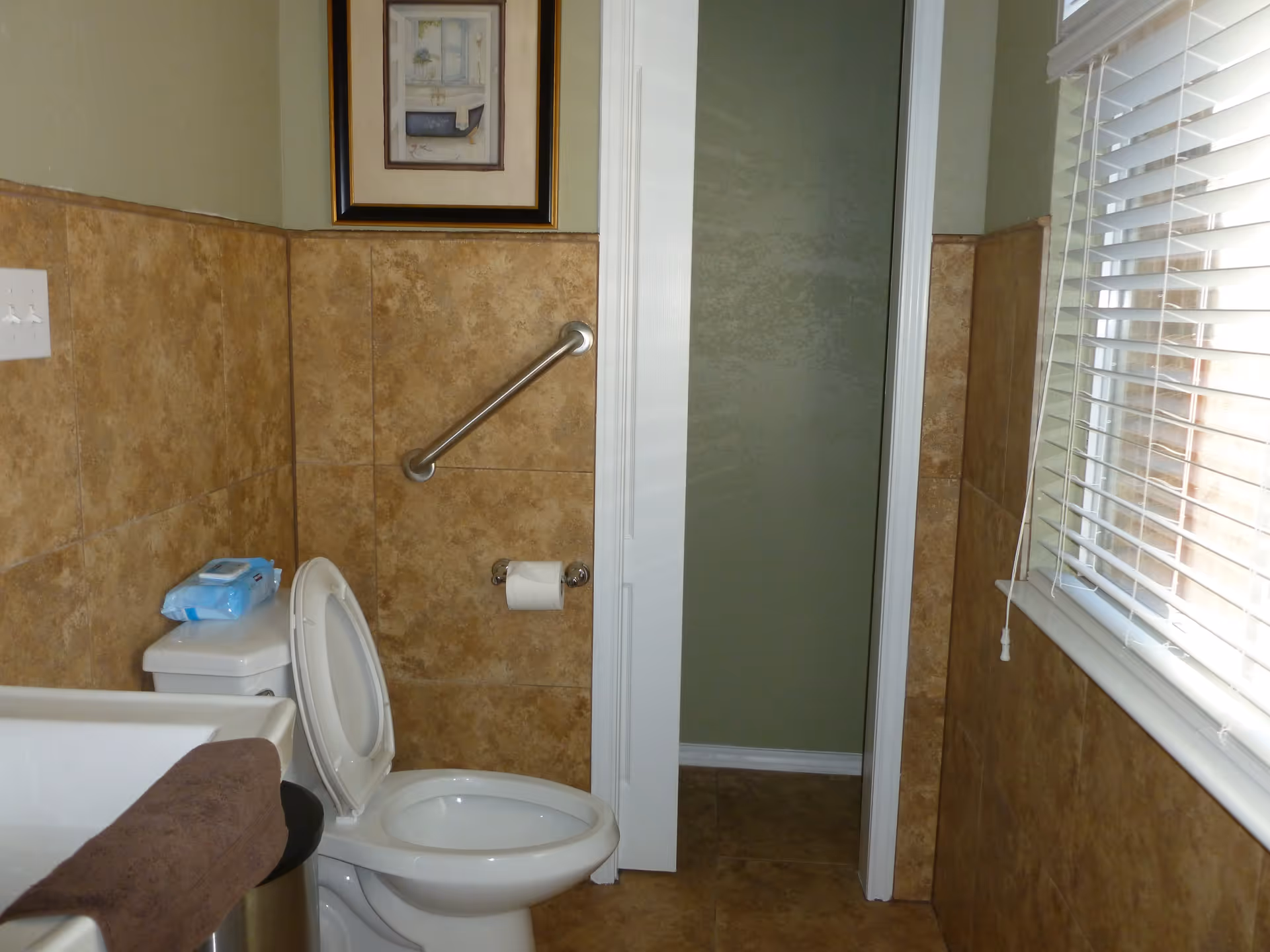 A bathroom with beige tiled walls and floor, featuring a white toilet with the lid open, a toilet paper holder with a roll of toilet paper, a grab bar on the wall, a framed picture above the grab bar, a window with white blinds, and a white door slightly ajar.