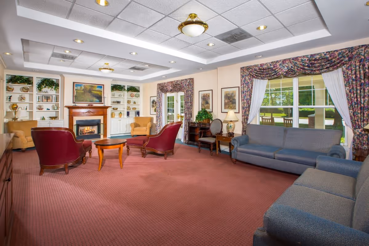A spacious living room with red carpet, two blue sofas, two red armchairs, and two yellow armchairs arranged around a wooden coffee table. The room features a fireplace with a painting above it, built-in white shelves with decorative items, large windows with floral curtains, and a ceiling with recessed lighting and a central light fixture.