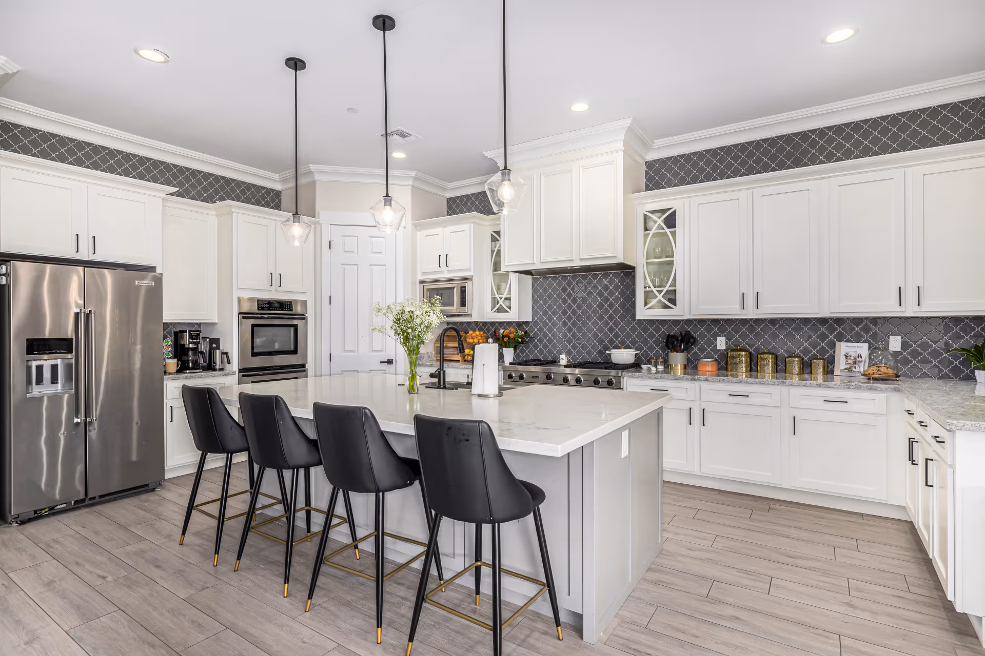 Modern kitchen with white cabinetry, a large island with a white countertop, four black bar stools, stainless steel refrigerator, built-in oven, and pendant lights hanging from the ceiling. The backsplash features a dark gray patterned tile, and the floor is light wood.