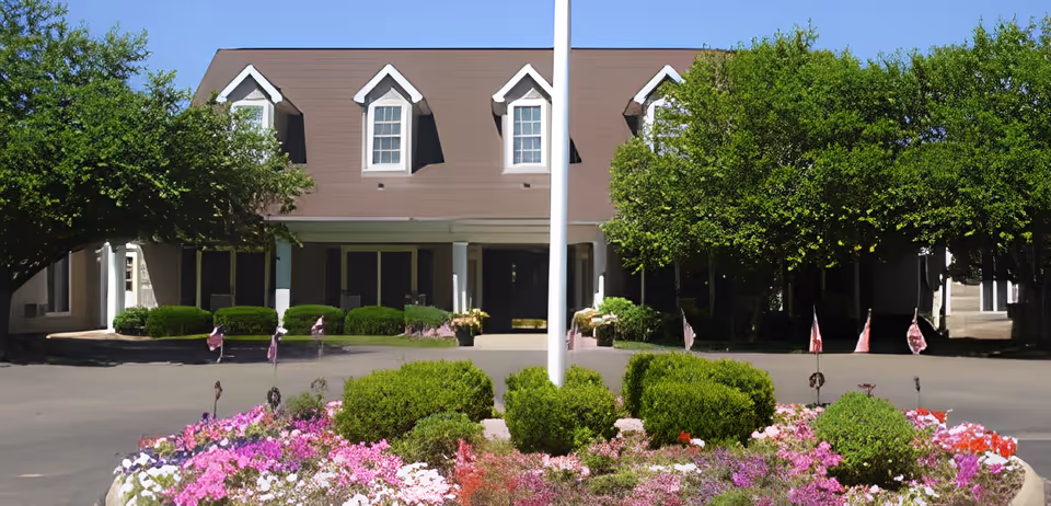 Front exterior view of a senior living facility with a brown roof, white dormer windows, and a landscaped garden with colorful flowers and green bushes in the foreground. There is a flagpole in the center and trees on either side of the building entrance.
