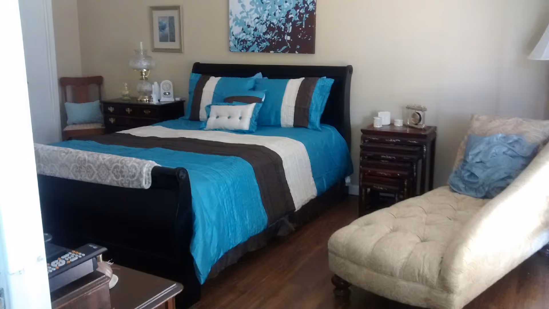 A bedroom with a black wooden bed frame and a blue, brown, and white striped bedspread. There are multiple pillows on the bed. To the right of the bed is a beige tufted chaise lounge with a blue pillow. Two dark wooden nightstands flank the bed, one with a lamp and the other with decorative items. The room has wooden flooring and light-colored walls with framed artwork above the bed.