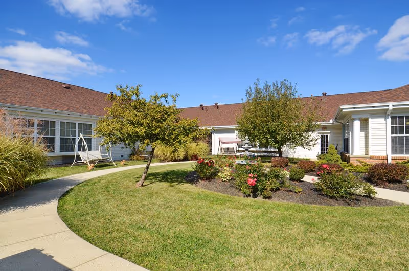 Sunny landscaped courtyard with a circular walkway, flower beds, small trees, and a single-story senior living building surrounding it.