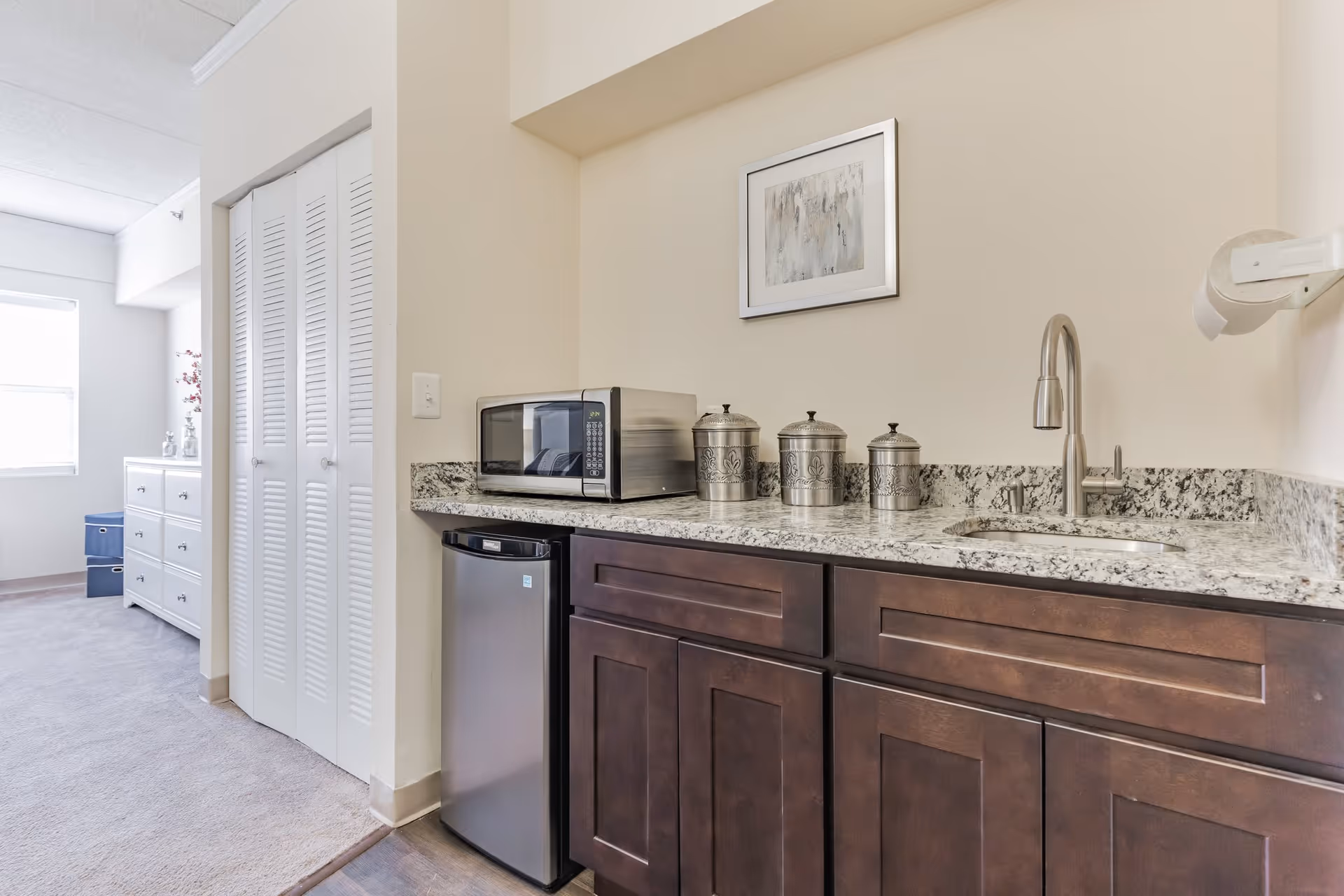 A kitchenette area with dark wooden cabinets, a granite countertop, a stainless steel sink with a modern faucet, a small stainless steel refrigerator, a microwave, and three decorative metal canisters. In the background, there is a white dresser and a window letting in natural light.