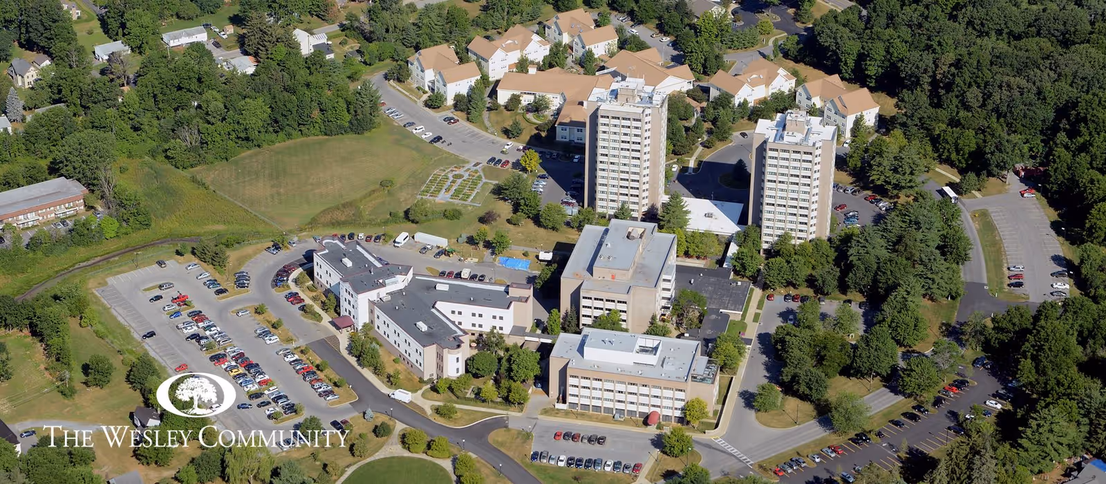 Aerial view of The Wesley Community - Embury Apartments showing multiple residential buildings surrounded by trees and parking lots. The complex includes several mid-rise apartment buildings, smaller housing units, and landscaped green areas with roads and parking spaces connecting the buildings.