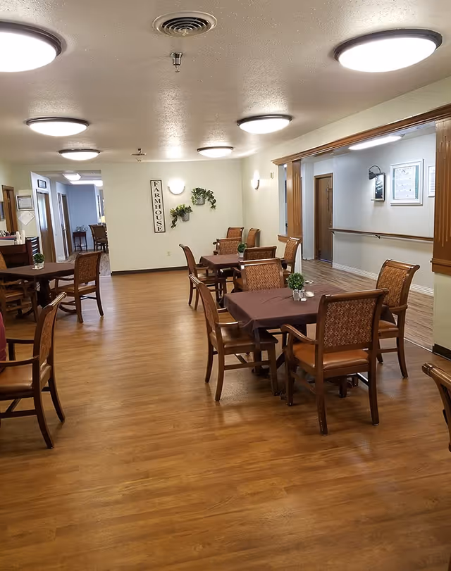 Dining room in a senior living facility with multiple tables and chairs on wood flooring under round ceiling lights.