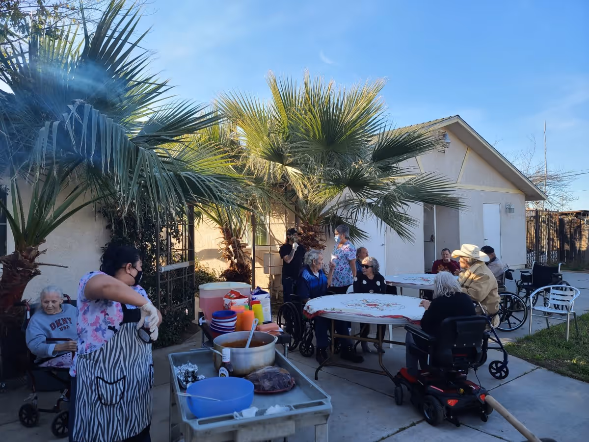Outdoor patio area at Rose Garden Assisted Living with several elderly residents sitting around tables covered with white and red tablecloths. Some residents are in wheelchairs and one is in a motorized scooter. A caregiver wearing a floral scrub top and face mask is preparing food at a cart with bowls and containers. Palm trees and a building with a white exterior are visible in the background under a clear blue sky.