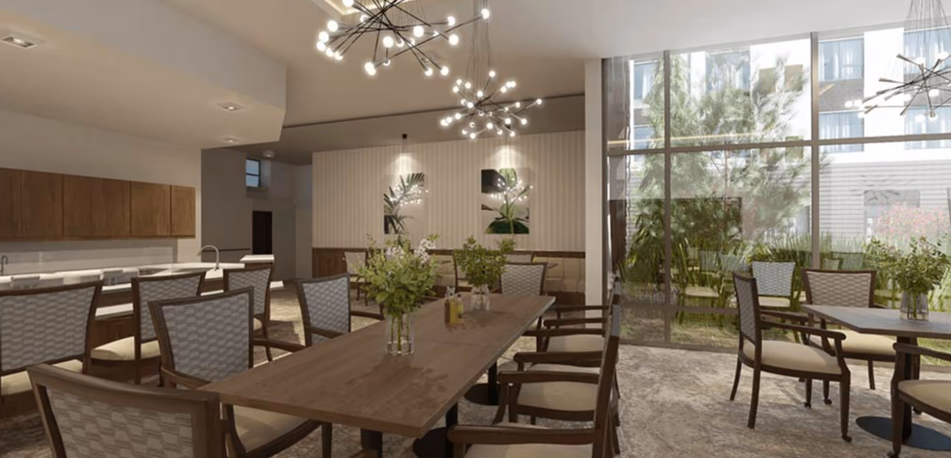 Bright communal dining room with a long wooden table, multiple chairs, modern chandeliers, and floor-to-ceiling windows overlooking greenery.