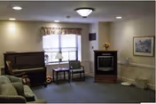 A cozy living room area with a piano on the left, two chairs and a small table in front of a window with a valance, a fireplace with a flower vase on top, a framed picture on the wall, and a couch partially visible on the left side.
