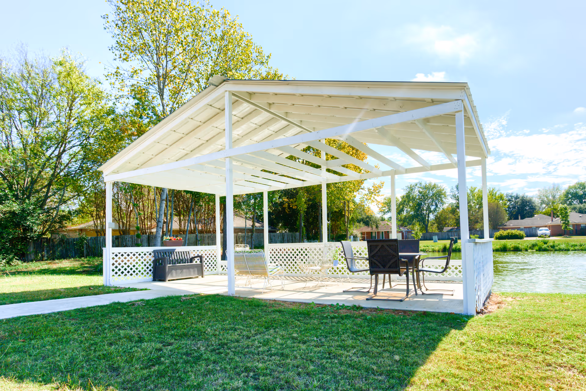 A white wooden gazebo with a slanted roof situated on a grassy area near a pond. The gazebo contains outdoor furniture including a bench, a swing, and a table with chairs. Trees and houses are visible in the background under a partly cloudy sky.