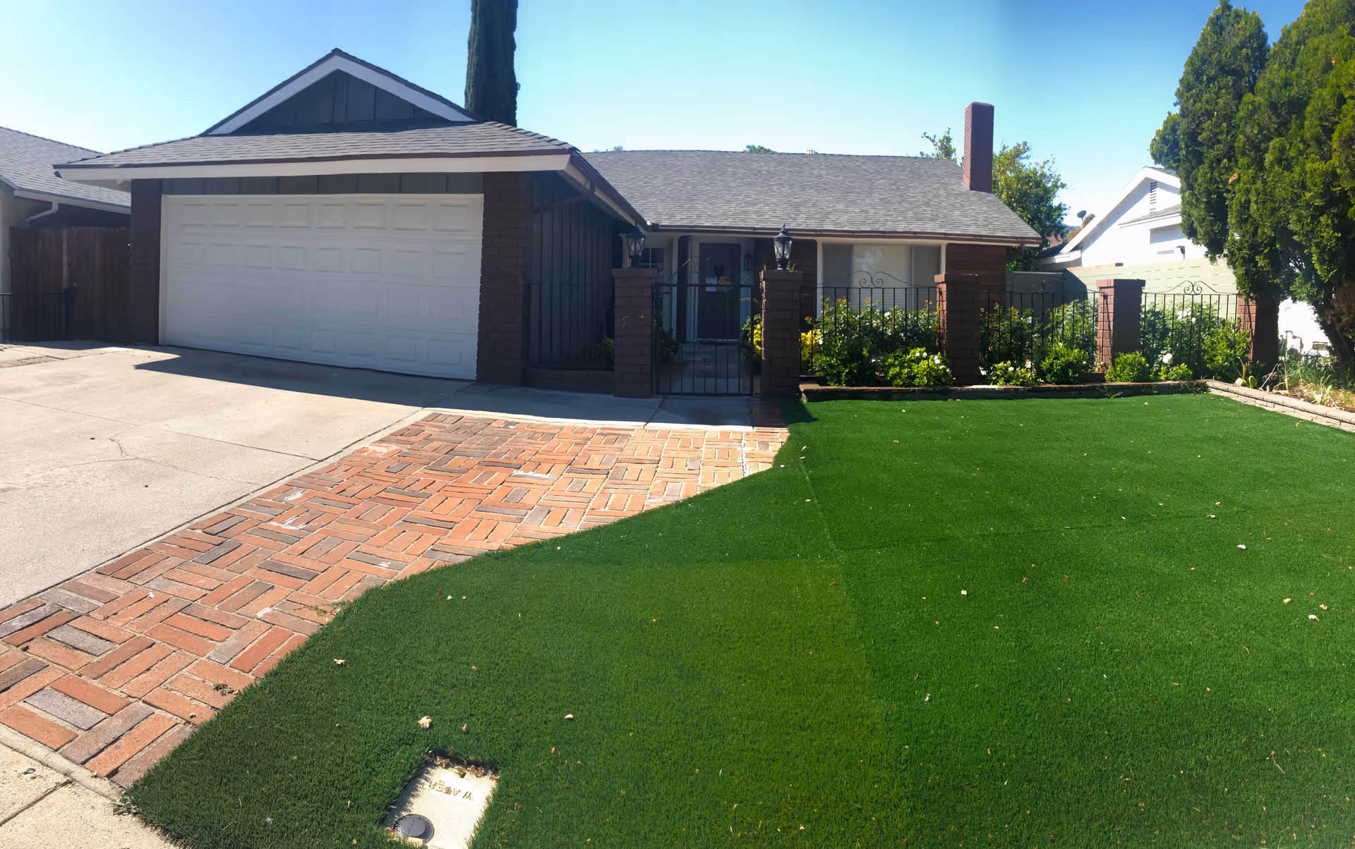 Single-story house front with an attached two-car garage, gated entrance, brick driveway and green lawn.