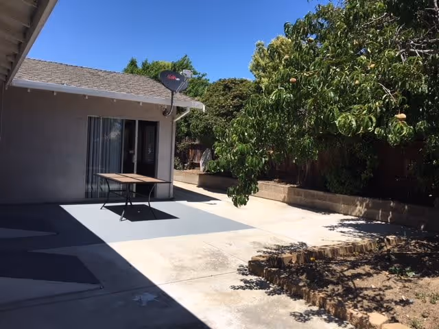 Outdoor patio area with a concrete floor, a small table, a sliding glass door leading into the building, a satellite dish mounted on the roof, and a tree with green leaves and some fruit. There is a garden bed with soil and a stone border on the right side.