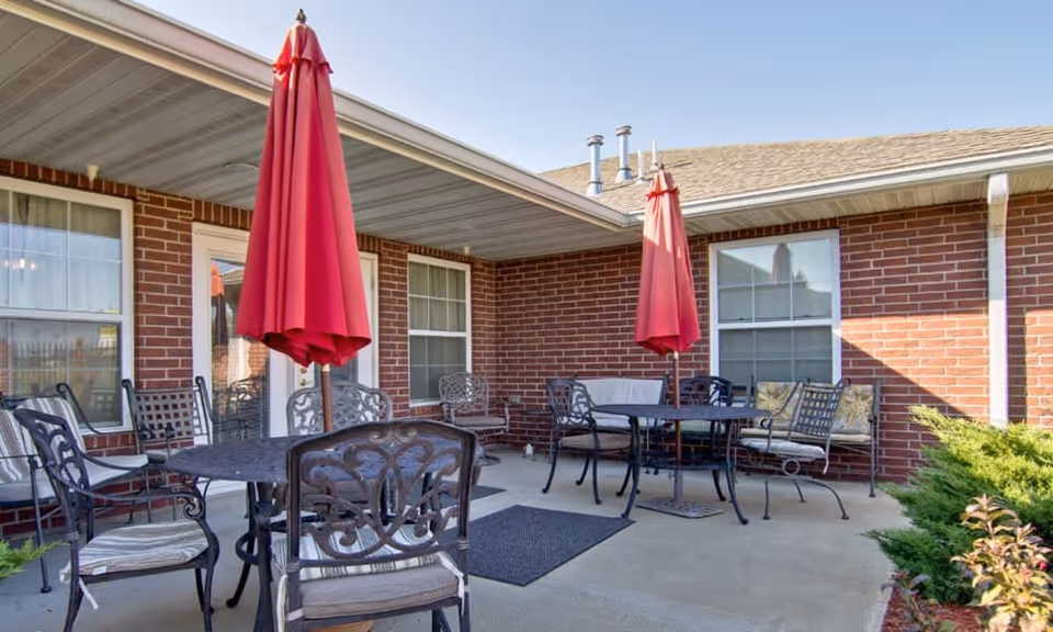 Outdoor patio area at Silver Creek Senior Living with metal tables and chairs, two red closed umbrellas, and brick walls of the building in the background under a clear sky.