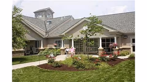 Front exterior of a single-story senior living building with landscaped flower beds, a walkway and outdoor seating on a sunny day.