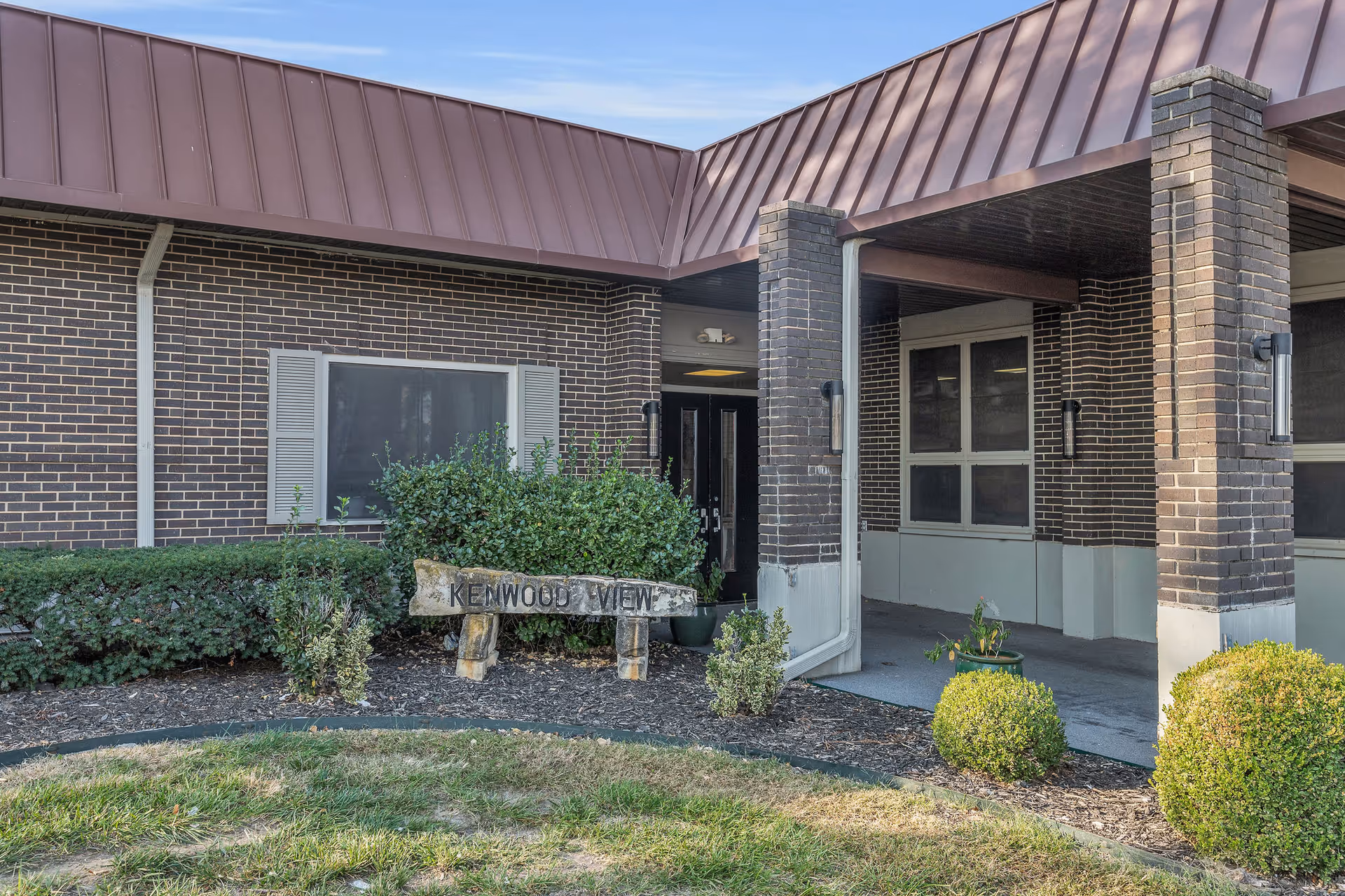 Exterior view of Kenwood View Health and Rehabilitation Center showing a brick building with a covered entrance, bushes, and a wooden sign with the facility name.
