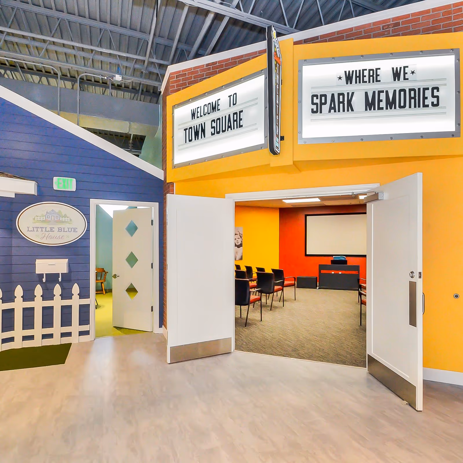 Interior view of a senior living facility area with a small blue house labeled 'Little Blue House' on the left and an open doorway leading to a room with chairs and a projector screen. Above the doorway, two signs read 'Welcome to Town Square' and 'Where We Spark Memories'.