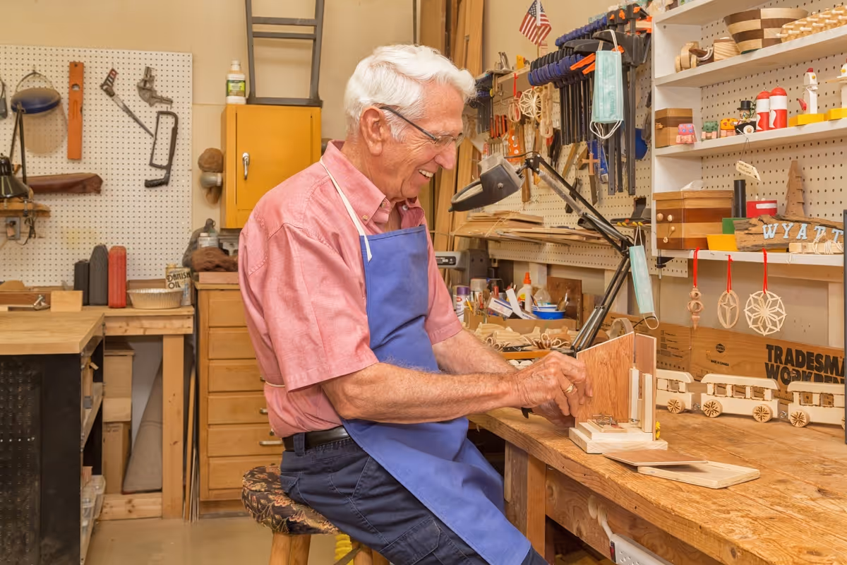 An elderly man wearing glasses and a blue apron is working on a woodworking project at a workbench in a workshop filled with tools, wooden crafts, and supplies.
