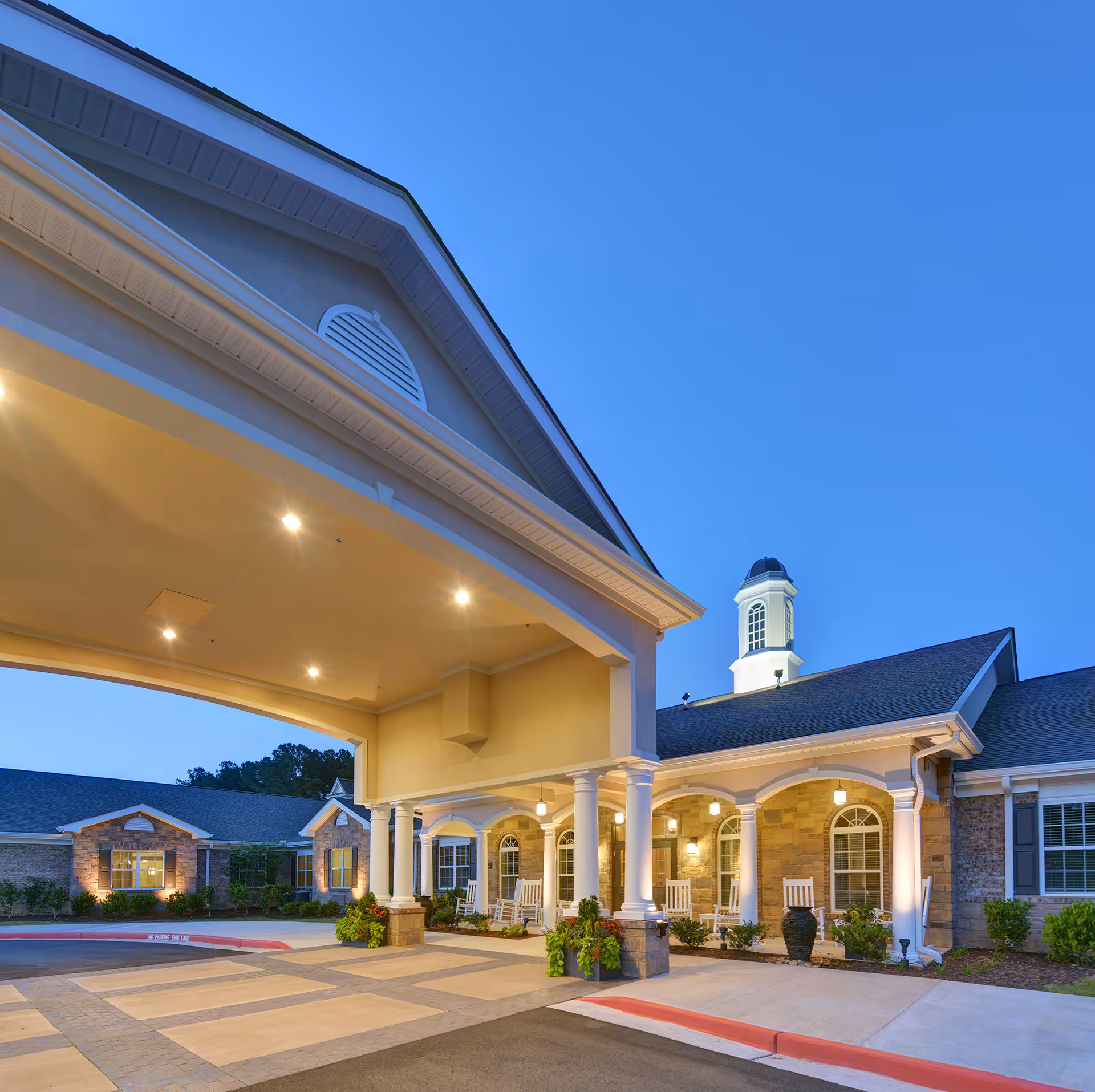Entrance of Benton Manor Of Stockbridge facility at dusk with a covered drop-off area supported by white columns, rocking chairs on the porch, and warm exterior lighting.