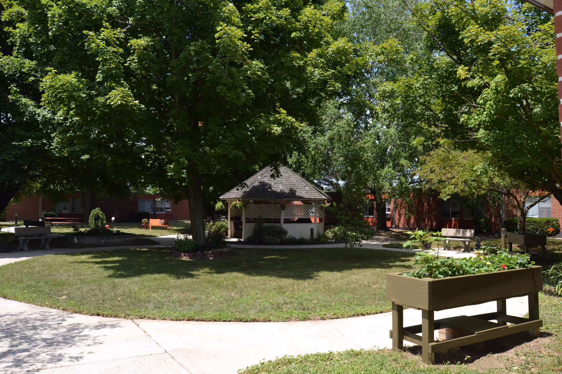 A peaceful outdoor courtyard area with a small gazebo surrounded by large green trees and well-maintained grass. There are benches and a raised garden bed with plants, and a paved walkway encircles the grassy area. The background shows parts of a brick building.