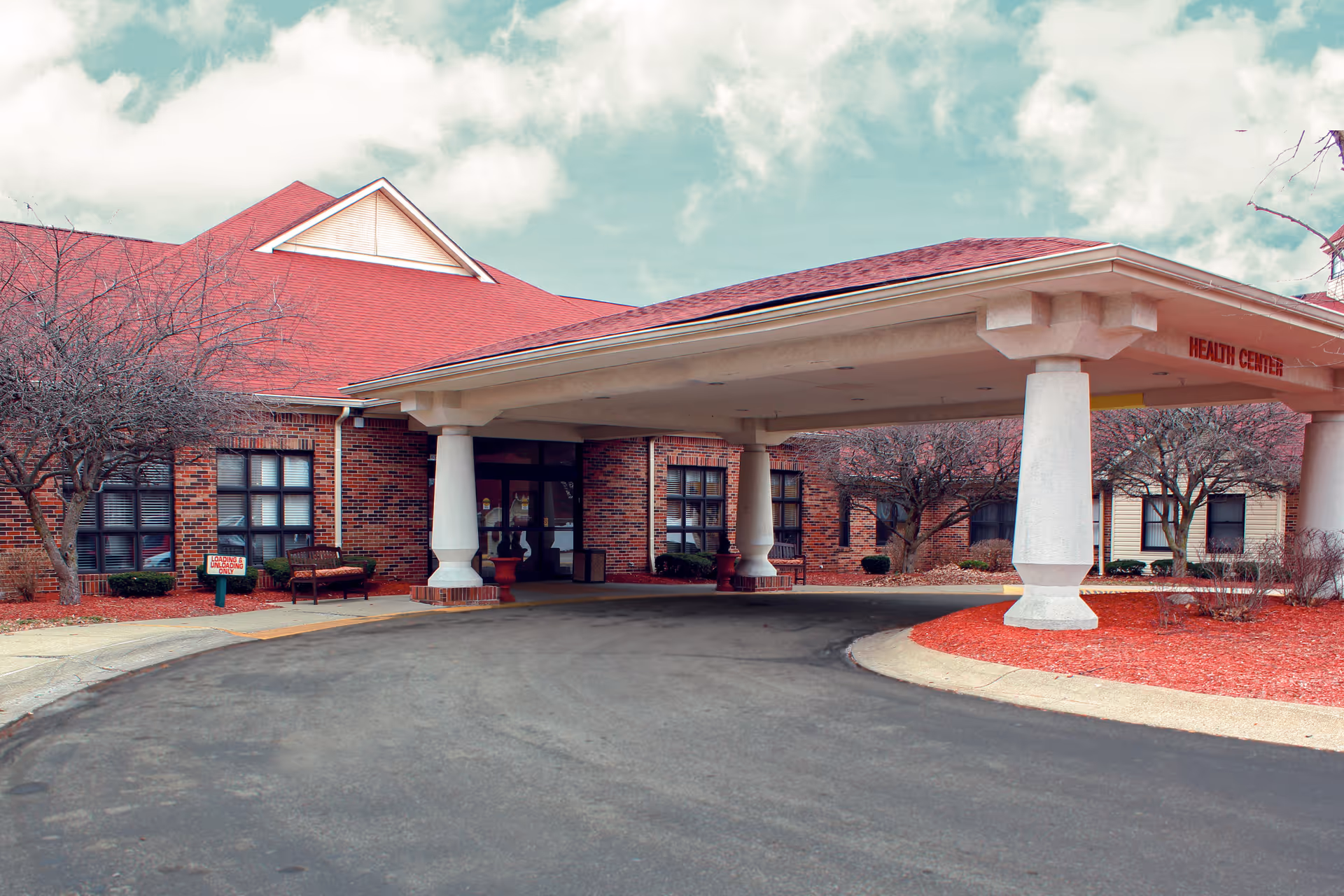 Entrance to a senior living facility with a covered driveway supported by large white columns. The building has red brick walls and a red roof. There are leafless trees and benches near the entrance, and a sign indicating 'Loading & Unloading Only'. The words 'Health Center' are visible on the covered driveway structure.