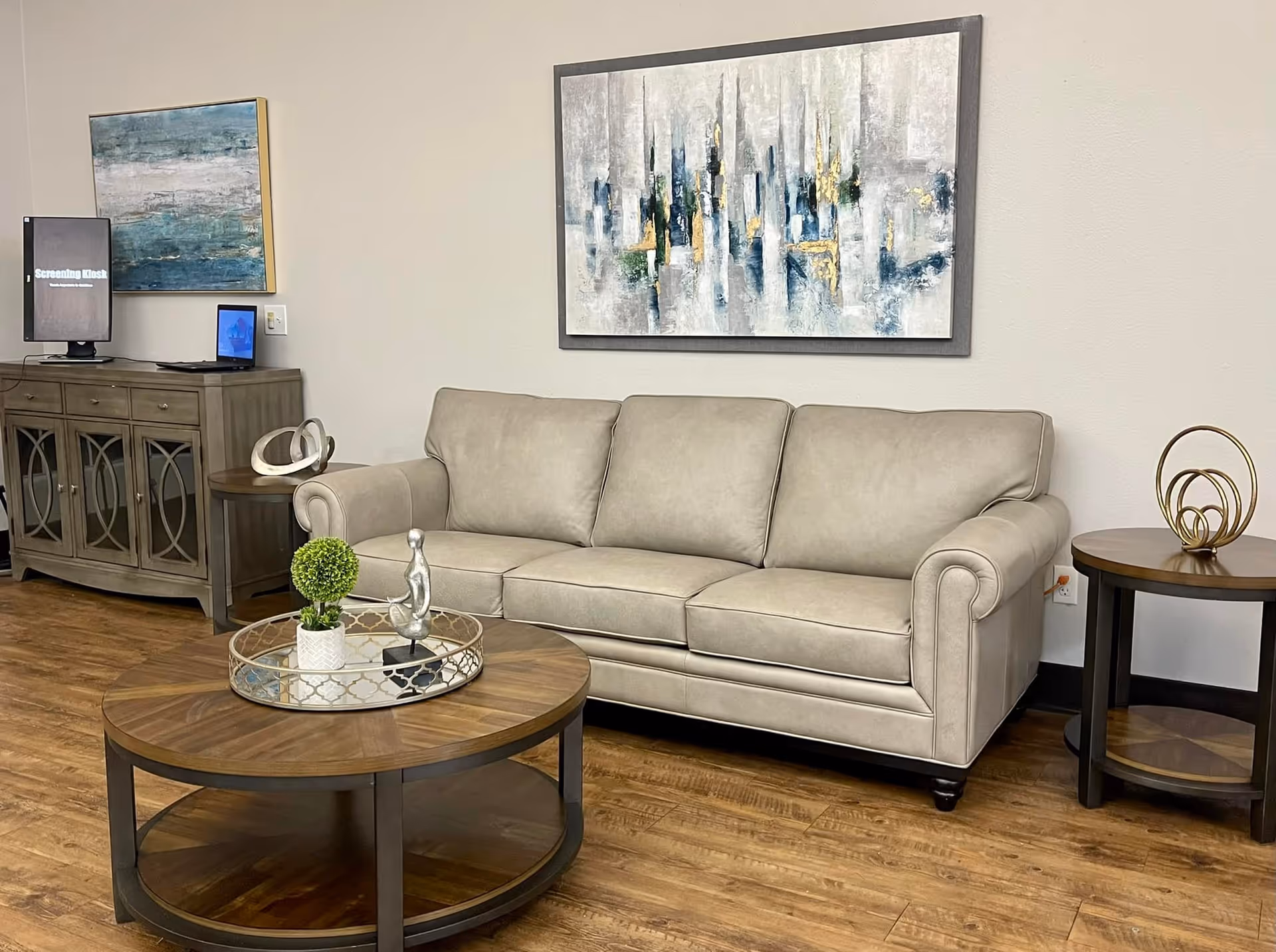 A cozy living room area with a beige three-seater sofa, a round wooden coffee table with decorative items including a small green plant and a silver figurine, and a matching round side table with a gold decorative sculpture. The room has wooden flooring and two abstract paintings on the wall. A wooden cabinet with a small screen labeled 'Screening Kiosk' is visible in the background.