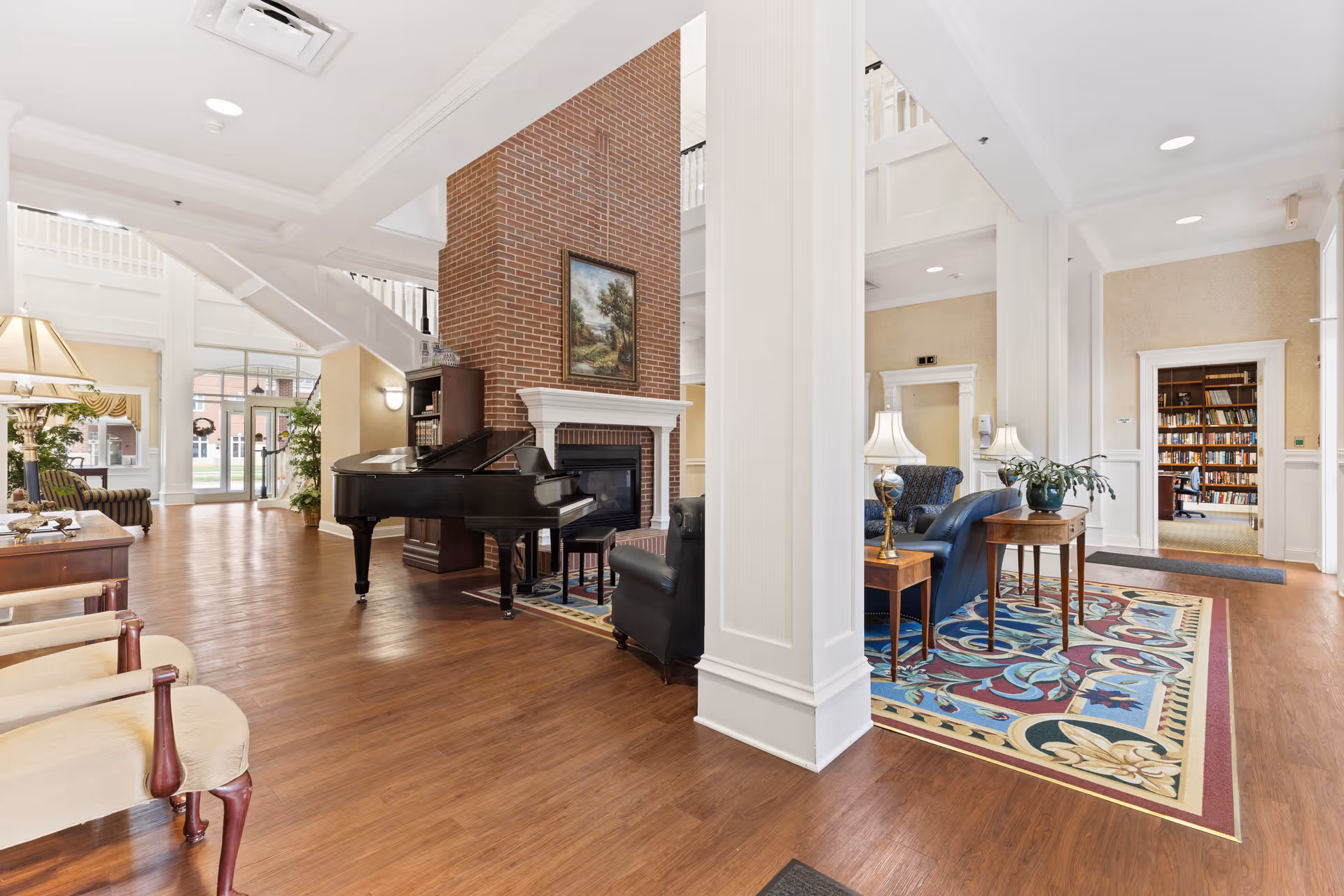 Spacious and well-lit senior living facility common area featuring a grand piano near a brick fireplace with a framed painting above it. The room has wooden flooring, comfortable seating including armchairs and sofas, side tables with lamps, and a colorful area rug. Large windows and glass doors allow natural light to fill the space, and a doorway leads to a room with bookshelves.