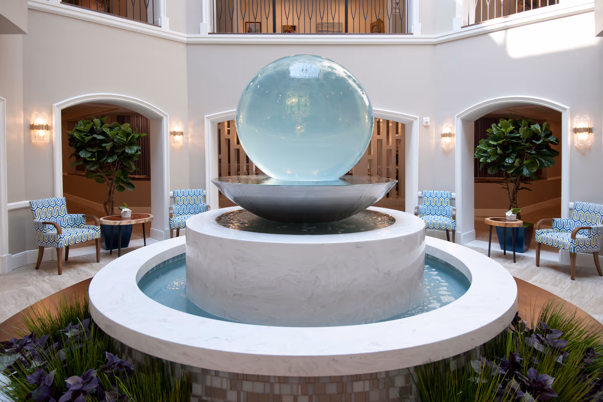 Indoor seating area with a large spherical water fountain in the center, surrounded by a circular marble basin. Four patterned armchairs and two small wooden tables with potted plants are arranged symmetrically around the fountain. The space features neutral walls, soft lighting from wall sconces, and large potted plants near arched doorways.
