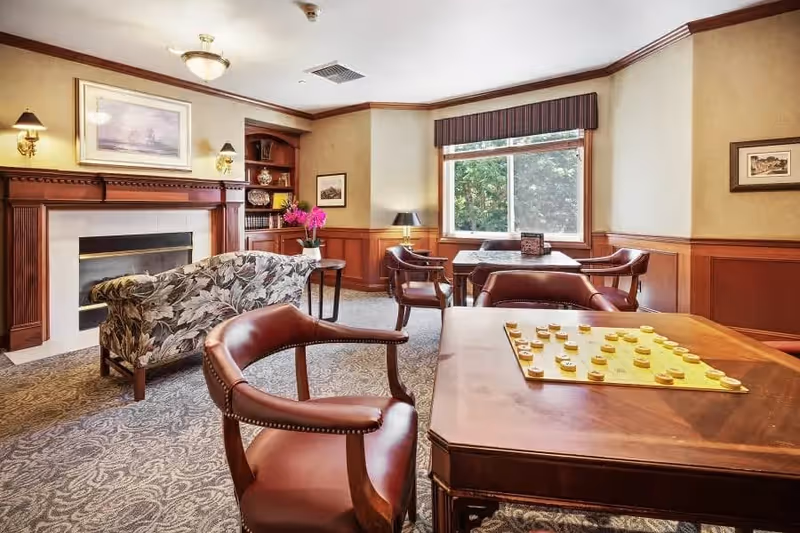 A cozy living room area with a floral patterned sofa facing a fireplace with a wooden mantle. There are several wooden tables and leather chairs arranged around the room, one table has a checkers game set up. The room has beige walls with wooden paneling, a large window with a valance, and decorative lighting fixtures on the walls.
