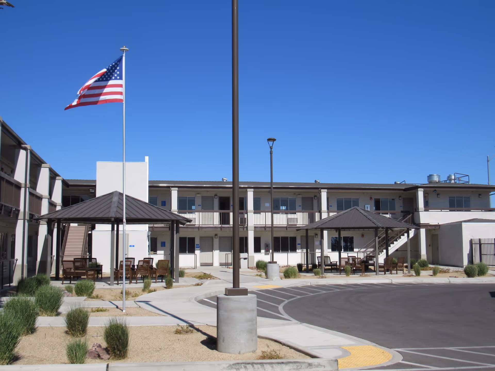 Outdoor courtyard area of a two-story senior living facility with beige buildings, two covered seating areas with benches and chairs, an American flag on a flagpole, and a clear blue sky.