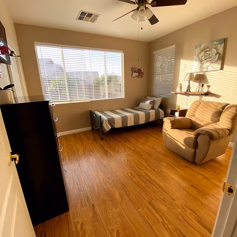 A bright bedroom with wooden flooring featuring a single bed with striped bedding, a beige armchair, a small nightstand with a lamp, a wall shelf with decorative items, and two large windows with white blinds letting in natural light.