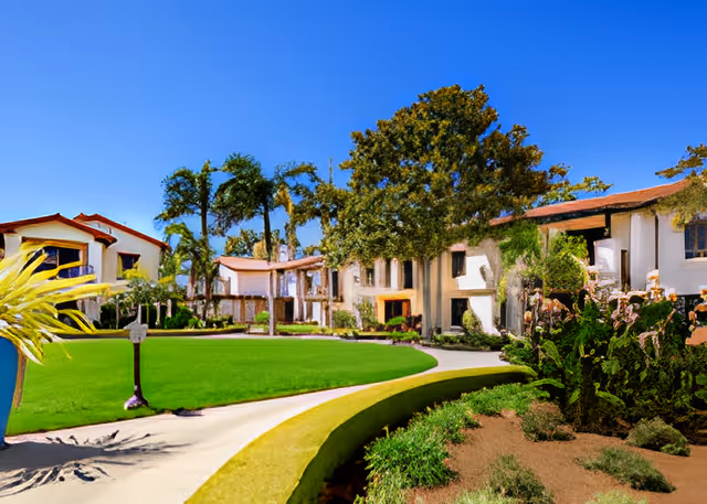 Sunlit courtyard and lawn in front of a Mediterranean-style senior living building with palm trees and walkways.