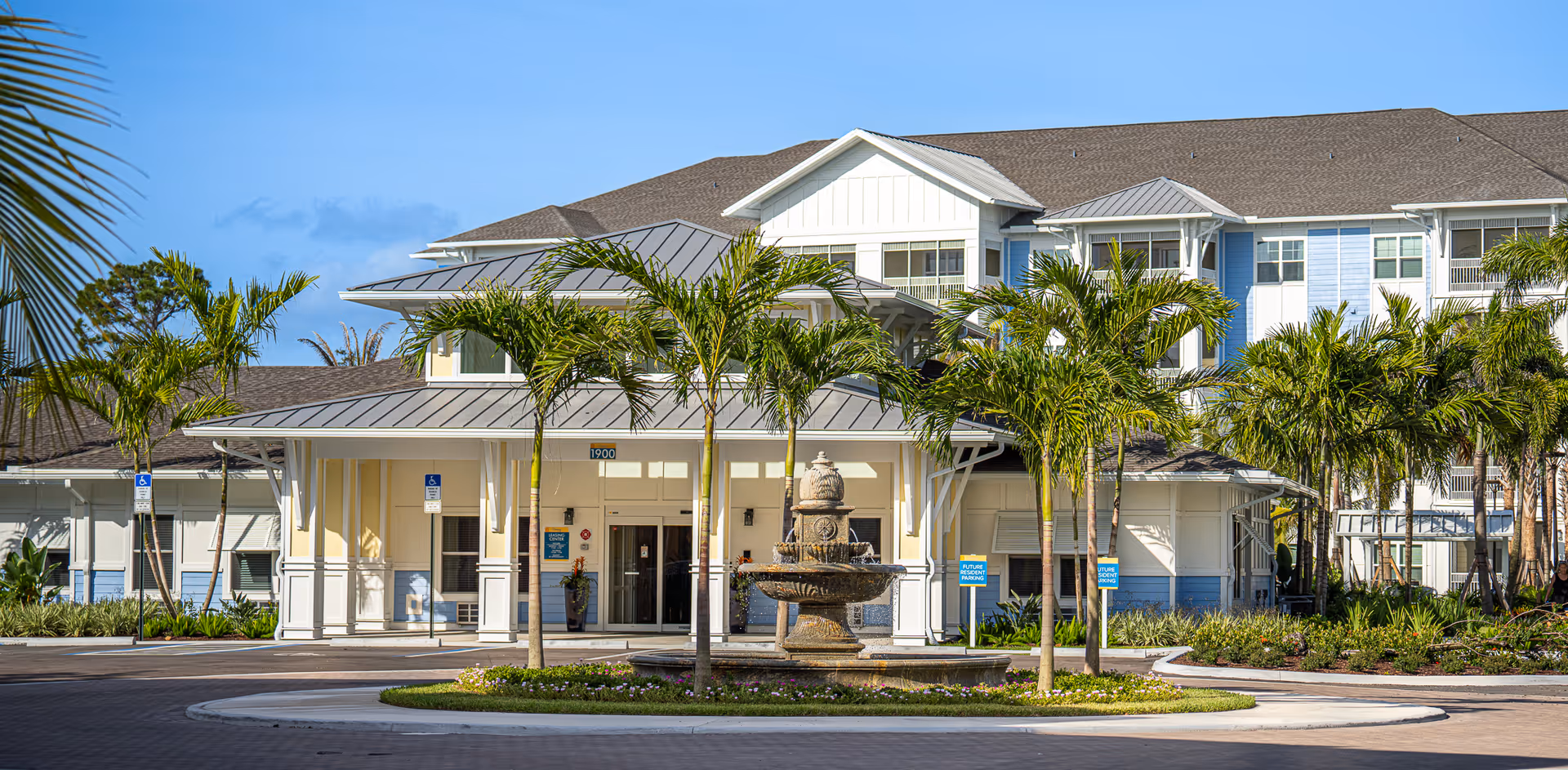 Front exterior view of The Sovana At Stuart facility with a circular driveway, palm trees, and a decorative fountain in the center. The building has a light blue and white facade with a gray roof under a clear blue sky.