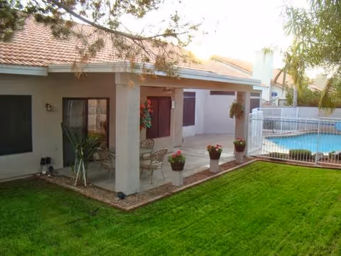 A backyard patio area of a residential building with a covered seating area featuring a table and chairs. There are potted plants along the edge of the patio, a well-maintained green lawn, and a fenced swimming pool in the background. Trees and shrubs surround the area.
