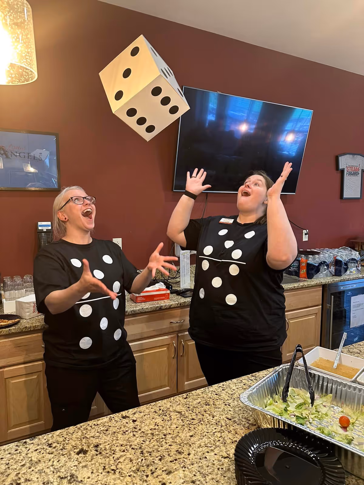 Two women wearing black shirts with white dice dot patterns are indoors, joyfully tossing a large foam dice in the air above a kitchen counter with food trays and utensils. The background shows a mounted flat-screen TV and kitchen cabinets.