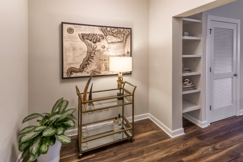 A stylish interior corner featuring a gold bar cart with a lamp and bottles, a framed map on the wall, a potted plant, built-in shelves, and a louvered door.