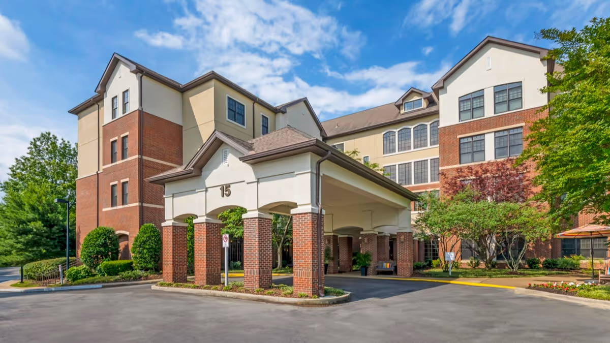 Exterior view of a multi-story senior living facility building with a covered entrance supported by brick columns, surrounded by trees and landscaping under a partly cloudy sky.