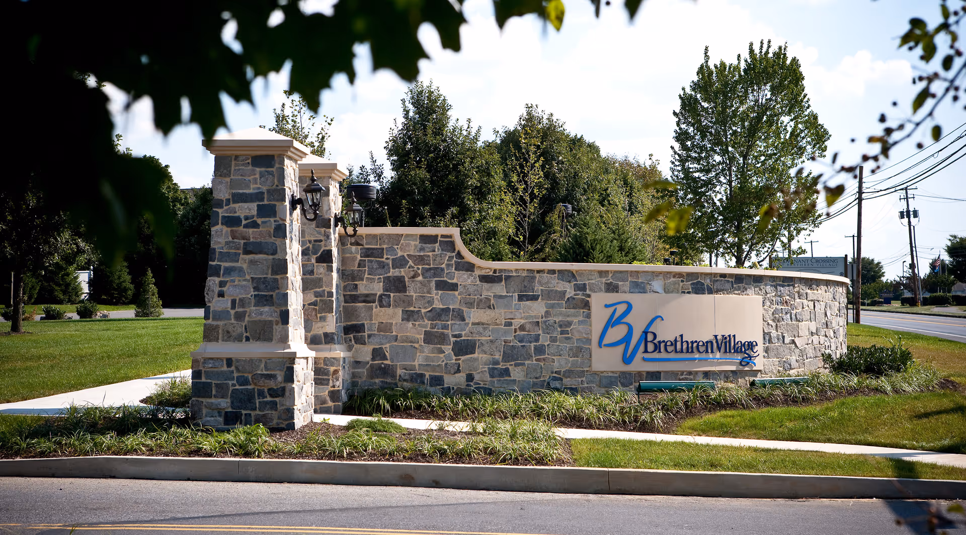 Stone entrance sign for Brethren Village with a curved stone wall and two stone pillars, surrounded by grass and landscaping, with trees and a road in the background.