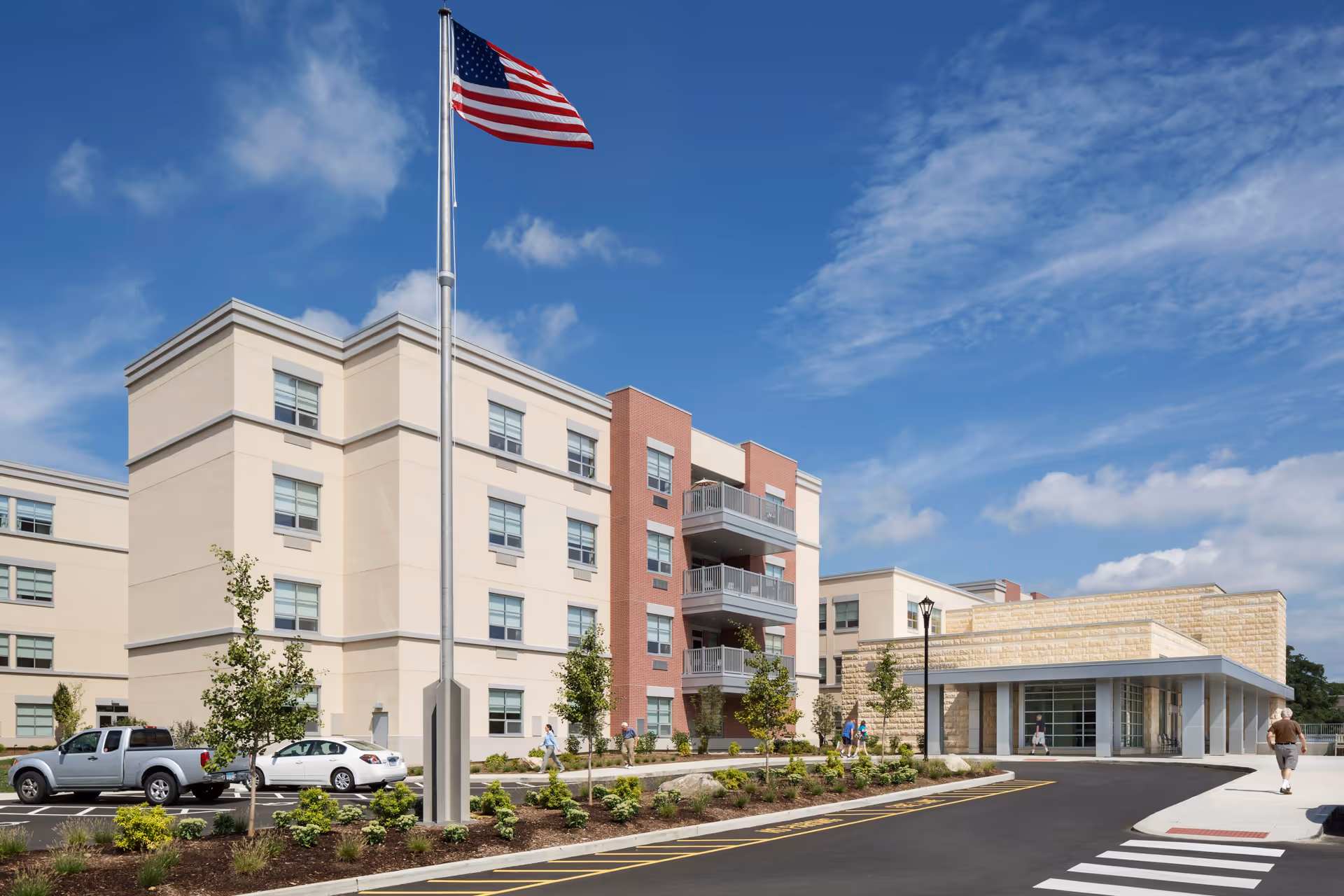 Exterior view of a multi-story senior living facility building with beige and red brick walls, balconies, a large American flag on a flagpole, a parking area with cars, landscaped greenery, and several people walking on the sidewalk under a partly cloudy blue sky.