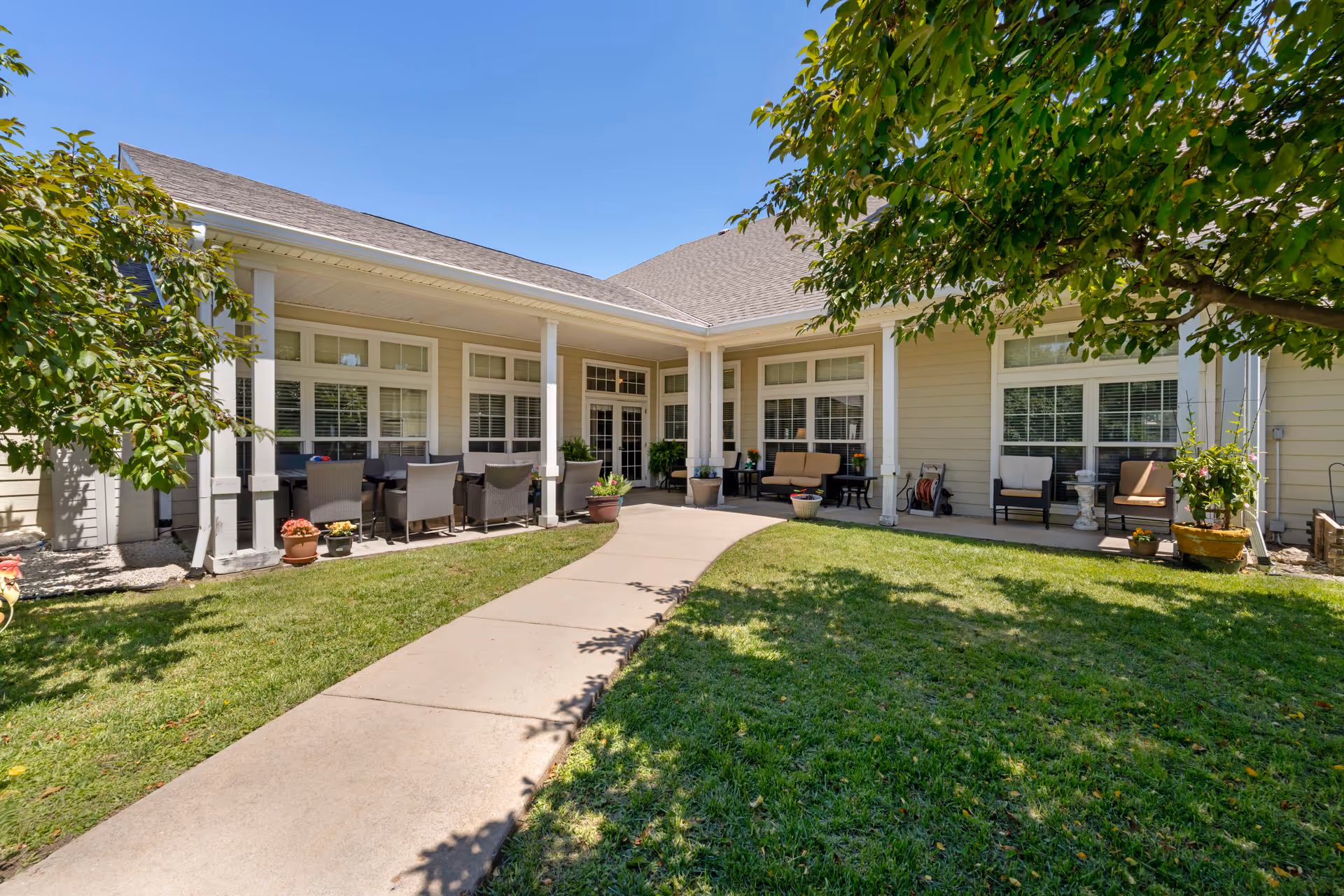 Outdoor patio area of a senior living facility with a concrete walkway leading to a covered seating area. The patio features multiple chairs, tables, potted plants, and greenery with trees providing shade. The building has large windows and a beige exterior under a clear blue sky.