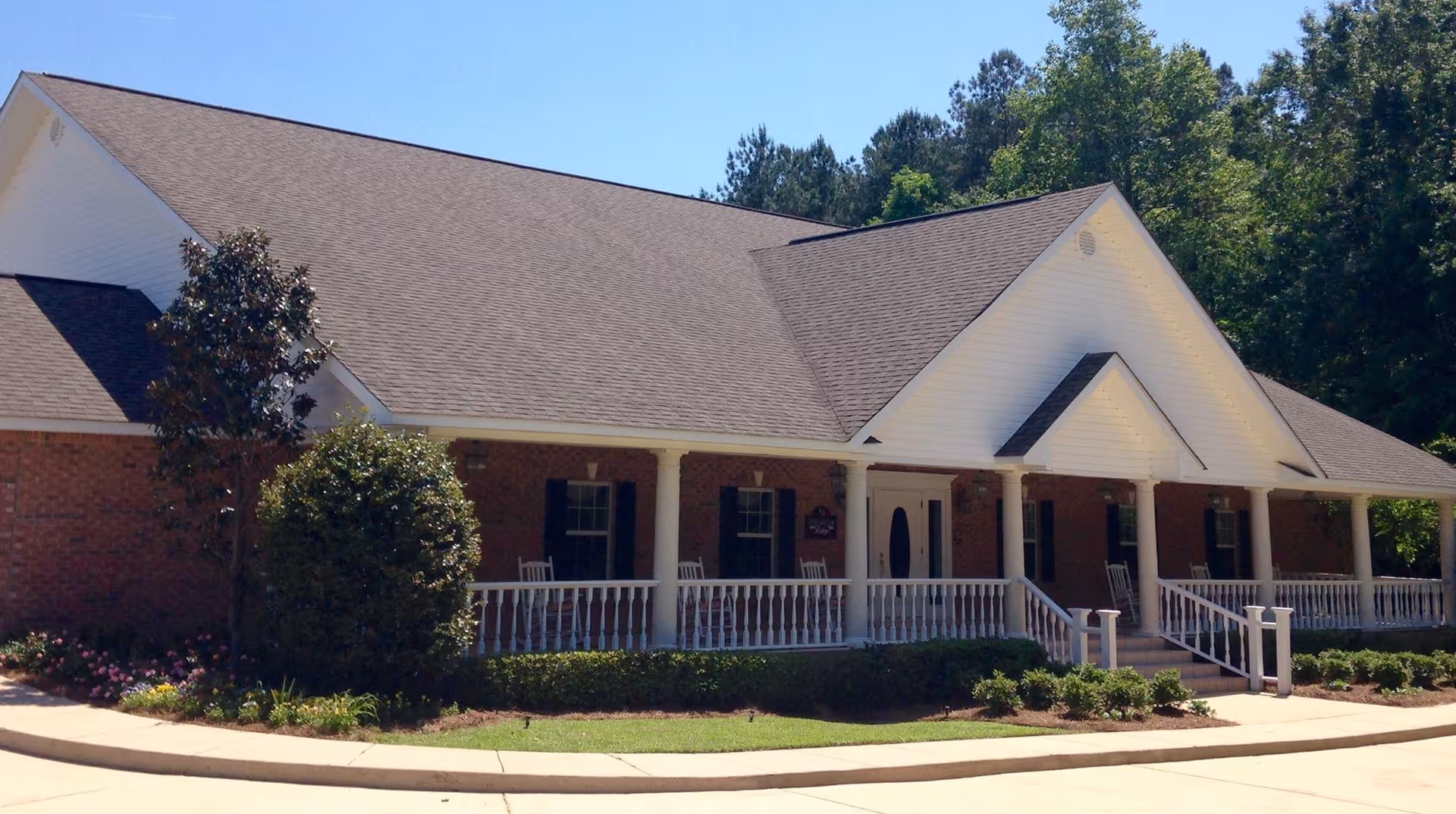 Exterior view of a single-story brick building with a large covered front porch featuring white railings and rocking chairs. The building is surrounded by neatly trimmed bushes and trees under a clear blue sky.