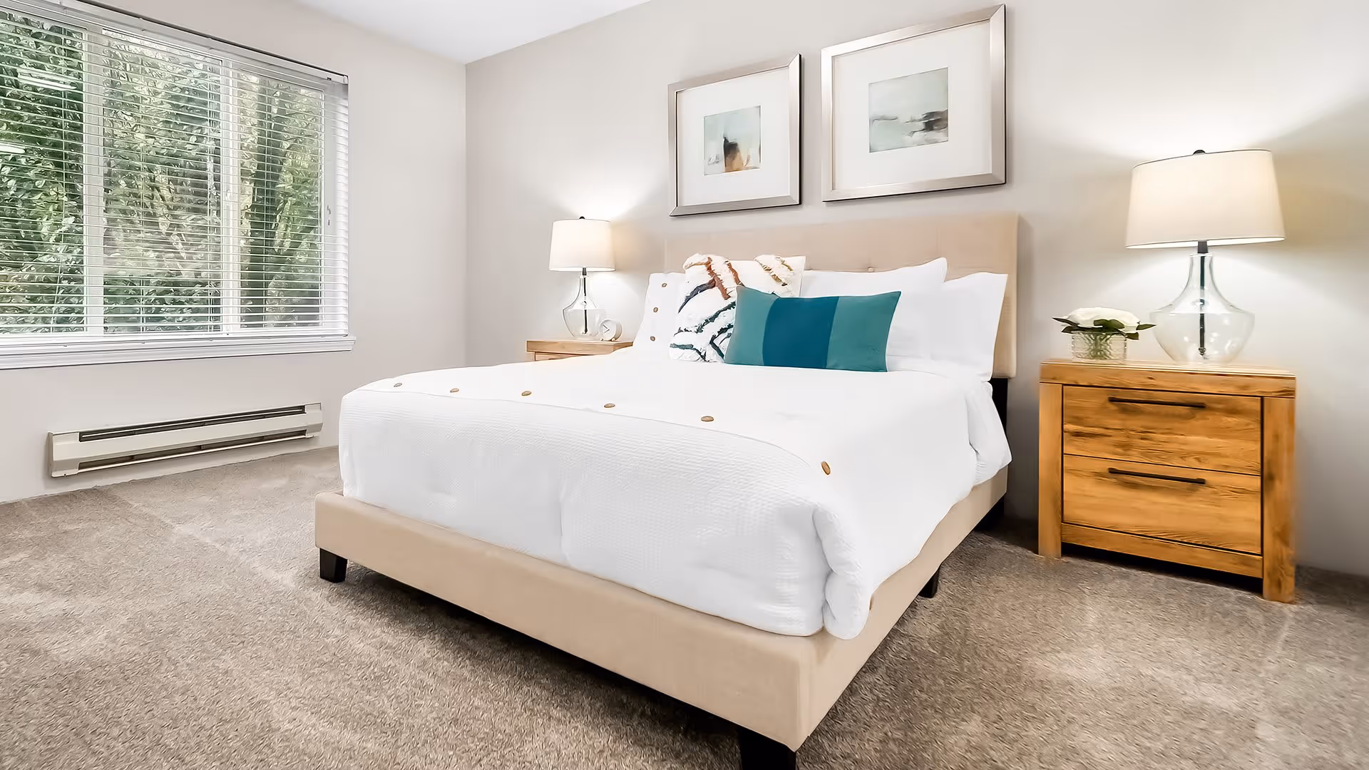A bright bedroom with a large beige upholstered bed dressed in white linens and decorative pillows. There are two wooden nightstands on either side of the bed, each with a glass-based lamp. Two framed abstract artworks hang above the bed. A window with white blinds lets in natural light, and the floor is carpeted in a neutral tone.